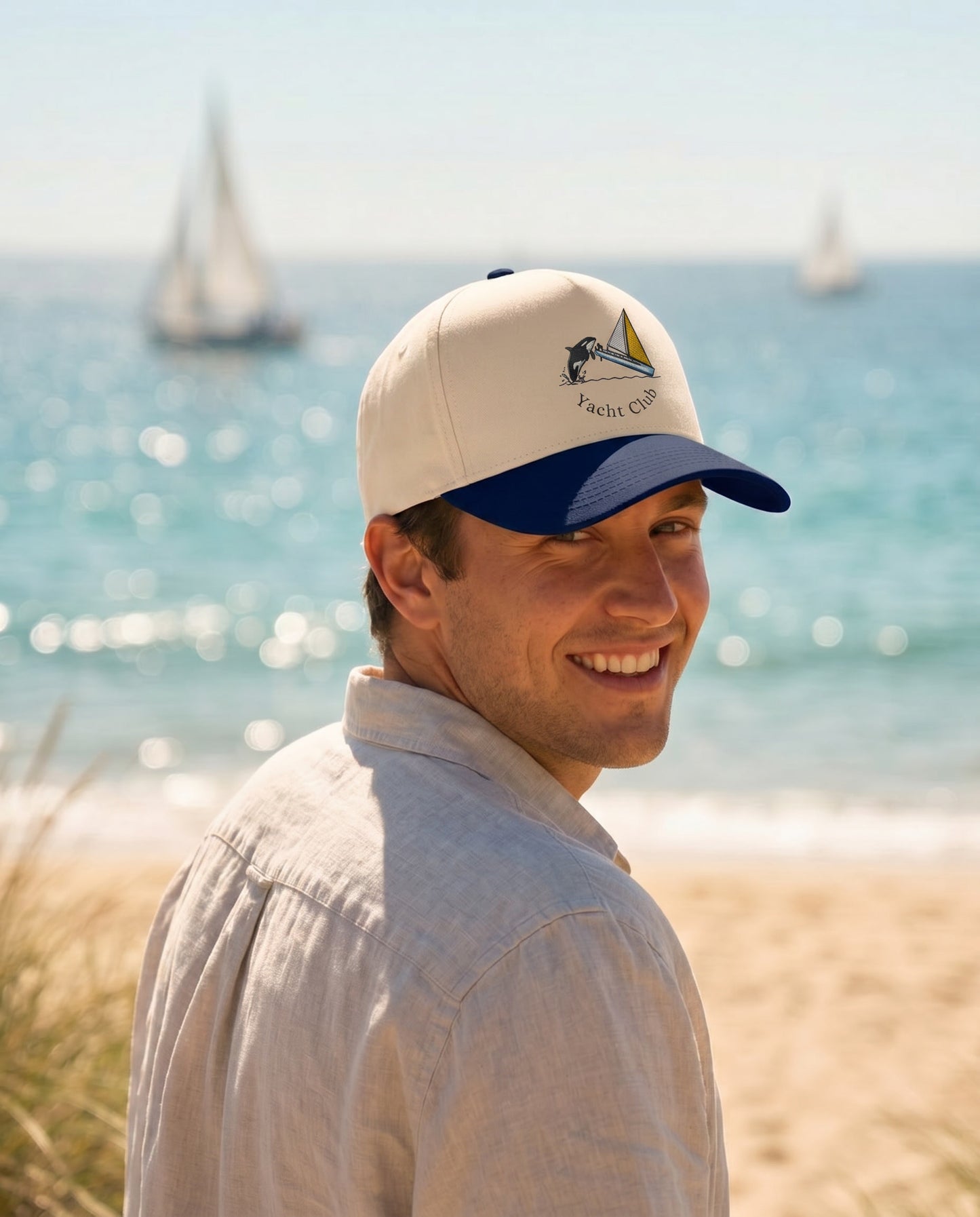 A man wearing a cream and navy blue baseball cap with a sailboat logo that reads 'FRESH SEA' stands on a beach with the ocean and sailboats blurred in the background.